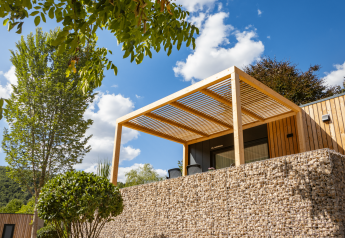 Lodge moderne Domus à Kohnenhof, Luxembourg, avec pergola en bois, mur en pierre et arbres sous ciel bleu.