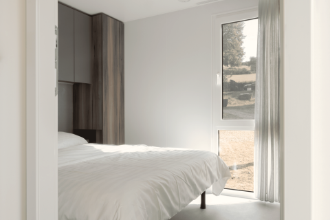 Lichte, moderne lodge slaapkamer met eenvoudig wit bed, grote ramen en uitzicht op de buitenomgeving.