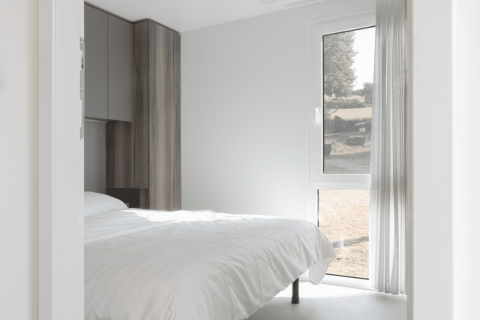 Habitación luminosa de una cabaña moderna con cama blanca sencilla, ventanal y armario de madera.