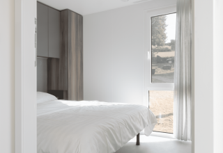 Chambre claire et moderne d’un lodge avec grand lit blanc, fenêtre panoramique et placard en bois.