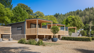 Lodge moderne Silvanus avec terrasse en bois, entouré de verdure et de forêt sous un ciel bleu clair.