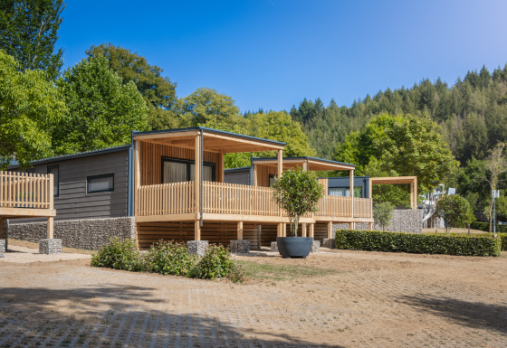 Lodge moderne Silvanus avec terrasse en bois, entouré de verdure et de forêt sous un ciel bleu clair.