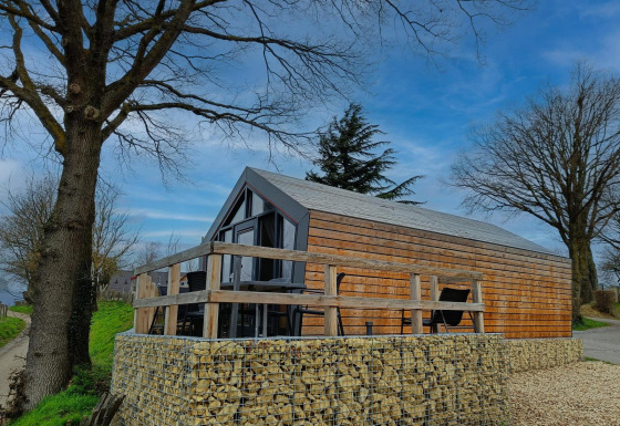 Solo Retreat lodge i træ med stor terrasse og stenmure, omgivet af store træer og blå himmel.