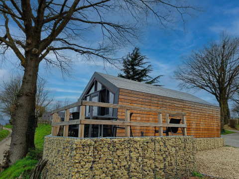 Solo Retreat lodge met houten buitenzijde, terras in hout en stenen muur, tussen bomen en blauwe lucht.