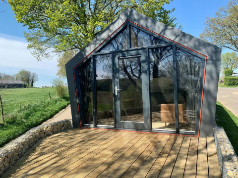 Et moderne Solo Retreat-lodge i Gulperberg, Holland, med glasfacade og træterrasse under træer.