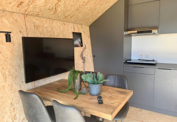 Interno di un lodge moderno con tavolo in legno, quattro sedie, TV a parete, cucina grigia e piante.