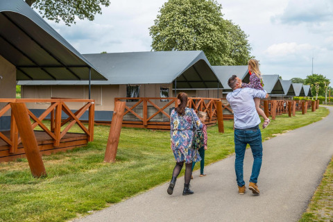 A family walks by luxury glamping tents with wooden decks at a scenic campground under leafy trees.