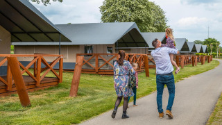 Una famiglia passeggia davanti a tende glamping di lusso con verande in legno in un campeggio alberato.