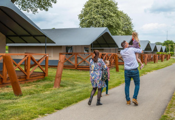 En familie går foran komfortable glampingtelte med træterrasser på en campingplads under trækroner.
