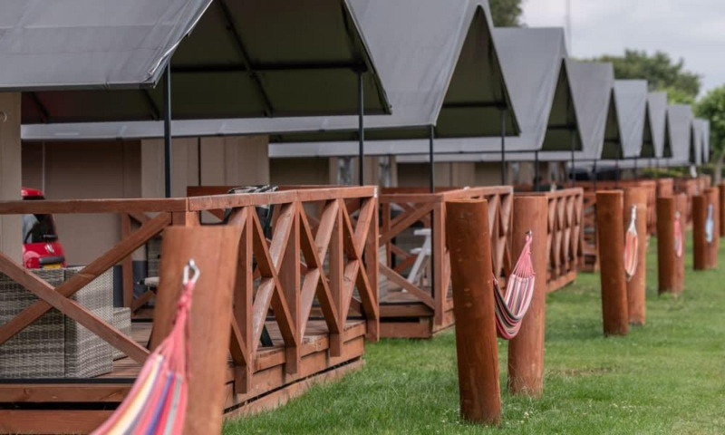 Fila de cabañas con terrazas de madera y hamacas en un césped verde, vista en un lodge bajo cielo nublado.