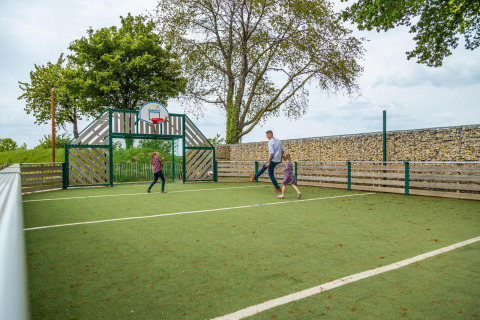 Familia juega fútbol en una pista al aire libre con canasta y árboles, en un alojamiento glamping.