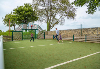 Gezin speelt voetbal op een buitensportveld met basketbalring bij een glamping, omgeven door bomen.