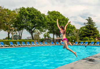 Une fille saute dans la piscine d'un hébergement glamping, entourée de transats et d’arbres verts.