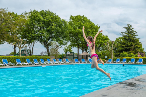 Una niña salta a la piscina en un alojamiento glamping, rodeada de tumbonas y árboles verdes.