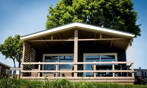 Un lodge moderne avec une terrasse couverte en bois, entouré d’arbres verts, sous un ciel bleu ensoleillé.