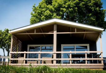 Een knusse lodge met overdekt terras, houten balustrade en zitjes, geflankeerd door groene zomerse bomen.