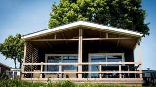Un lodge moderne avec une terrasse couverte en bois, entouré d’arbres verts, sous un ciel bleu ensoleillé.