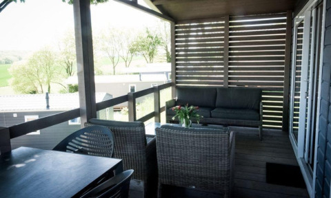 Terrasse couverte d'un lodge avec des meubles en rotin, vue sur des arbres et des mobil-homes autour.