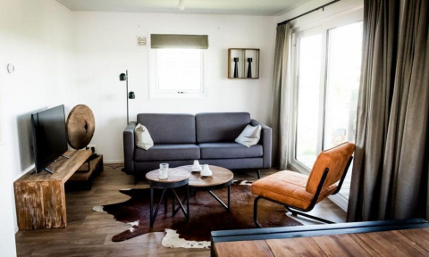 Salon lodge moderne avec canapé gris, fauteuil cuir, TV, table en bois rustique et grande fenêtre.