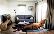 Stilfuld lodge-stue med grå sofa, brun læderstol, tv, bord og store vinduer, naturligt lys.