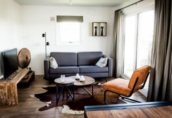 Stijlvolle lodge woonkamer met grijze zetel, leren stoel, TV, rustieke tafels en groot raam.