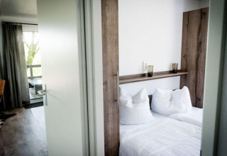 Camera lodge moderna con letto matrimoniale, biancheria bianca, grandi tende e poltrona in pelle.