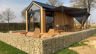 Een moderne Tiny House lodge met houten afwerking, stenen muur en zonnig terras met stoelen en hangmat.
