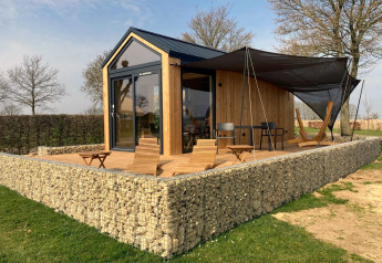 Een moderne Tiny House lodge met houten afwerking, stenen muur en zonnig terras met stoelen en hangmat.