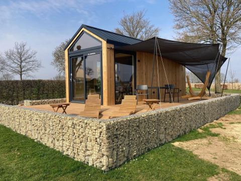 Moderna cabaña Tiny House con revestimiento de madera, muro de piedra y terraza con sillas y hamaca.