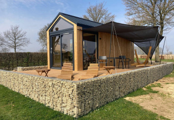 Moderna cabaña Tiny House con revestimiento de madera, muro de piedra y terraza con sillas y hamaca.