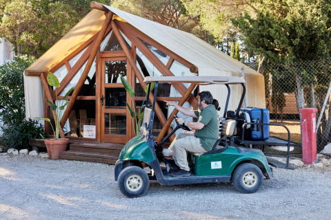 Hombre conduce carrito de golf con maletas frente a tienda glamping en TAIGA Conil, parque vacacional en Andalucía, España.