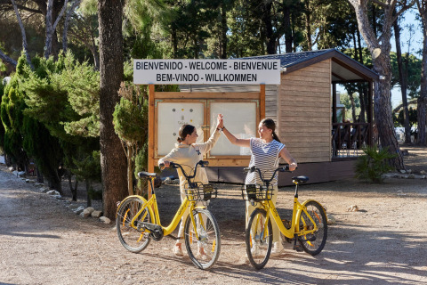 Zwei Frauen auf gelben Fahrrädern begrüßen sich vor dem Willkommensschild im TAIGA Conil Ferienpark, Andalusien.