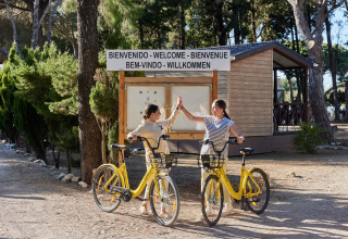Twee dames high-fiven op gele fietsen bij het welkomstbord aan TAIGA Conil vakantiepark in Andalusië, Spanje.