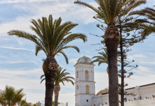 Palmer og hvidt tårn ved TAIGA Conil, en feriepark i Andalusien, Spanien, med blå himmel og skyer.