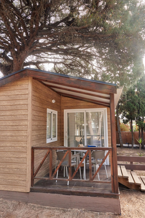 Bungalow Confort Poniente en TAIGA Conil, España, una acogedora cabaña de madera con porche bajo frondosos árboles.