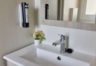 Baño moderno en Bungalow Confort Poniente de TAIGA Conil con lavabo, espejo, dispensador y maceta con flores.