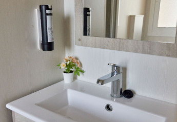 Baño moderno en Bungalow Confort Poniente de TAIGA Conil con lavabo, espejo, dispensador y maceta con flores.