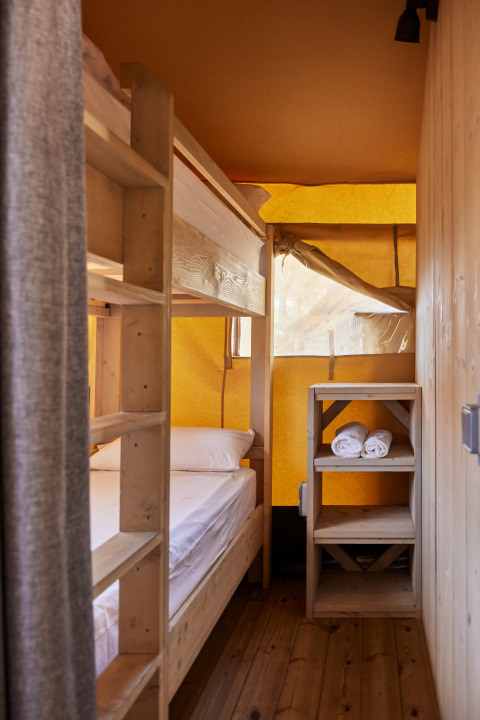 Interior of a safari tent featuring bunk beds, wooden shelves with rolled towels, and warm natural light.