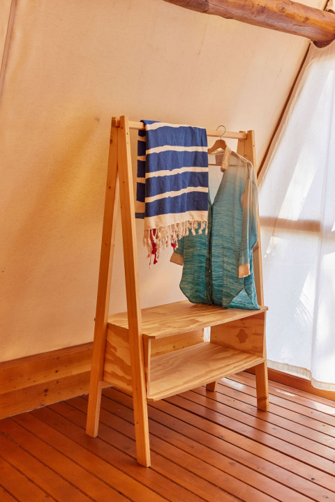 Træhylde med tøj og håndklæde i Glamping Tipi Almadraba, TAIGA Conil, Spanien, med sollys.