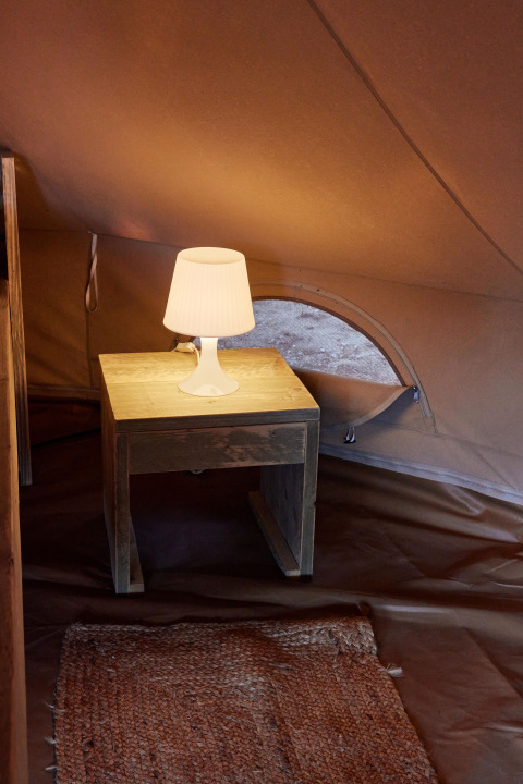 Interior de la tienda Glamping Bell Marisma en TAIGA Conil, España, con mesa de noche y lámpara encendida.