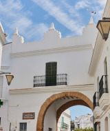 Vista de edificios blancos y un arco histórico en Conil de la Frontera, Andalucía, España, bajo un cielo azul.