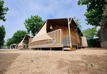 Safaritent Villatent Outback met houten veranda en luifel, omringd door bomen onder een heldere lucht.