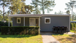 A cozy Easy chalet with a gray exterior, hedge and outdoor dining set, located in a peaceful forest area.