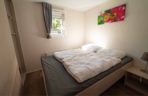 Schlafzimmer mit Doppelbett, weißer Bettdecke, Fenster, Schmetterlingsbild und Nachttisch im Easy chalet bei Eurocamping Vessem BV, Niederlande.
