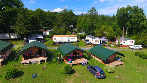 Foto de un camping con tiendas safari, caravanas y mucho césped verde, rodeado de árboles y naturaleza.