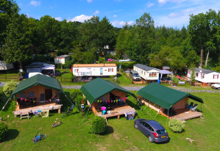 Foto de un camping con tiendas safari, caravanas y mucho césped verde, rodeado de árboles y naturaleza.