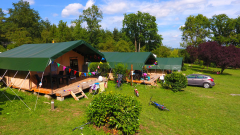 Safari-Zelte mit grünen Dächern auf einem Campingplatz, umgeben von Fahrrädern, Auto und Bäumen.
