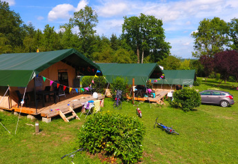 Safari tents with green roofs at a campsite, surrounded by bikes, a car, and lush green trees.