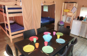 Interior de tienda safari con literas, mesa de comedor con platos de colores y zona de cocina equipada.