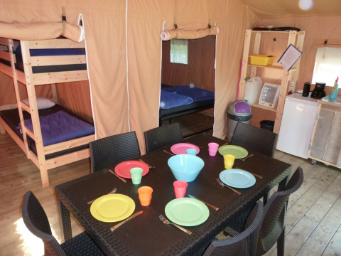 Binnenkant van een safaritent met stapelbedden, gedekte tafel met gekleurde borden en kleine kitchenette.